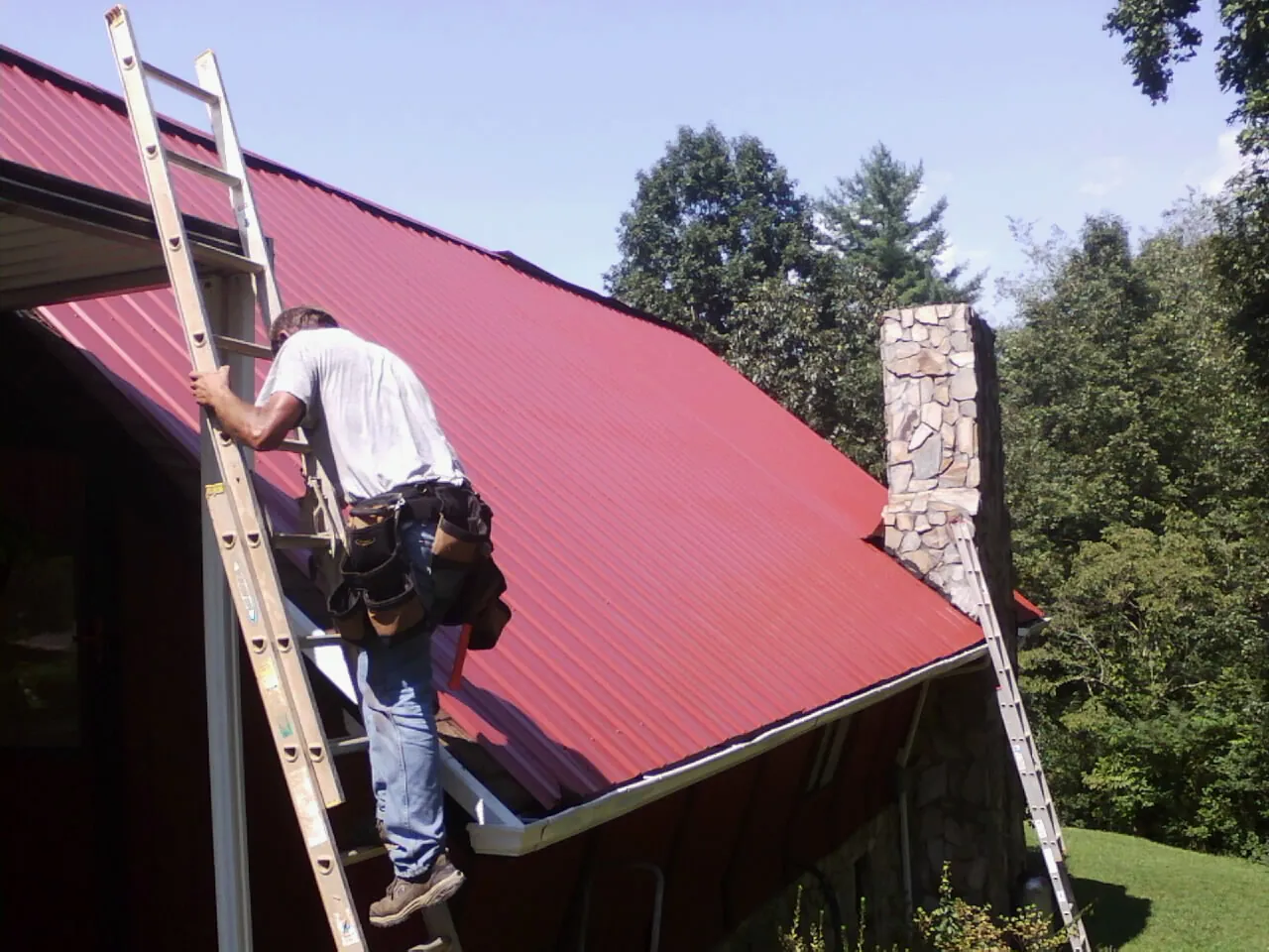 Roofer on a ladder beside a red metal roof and stone chimney — Soffit & Fascia Repair by Roofing Brothers in Center